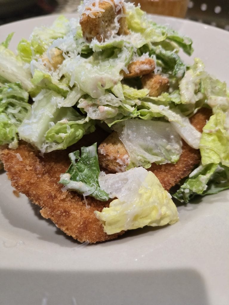 A close-up of a breaded and fried piece of meat, topped with chopped Caesar salad that includes romaine lettuce, croutons, and grated Parmesan cheese, all mixed with creamy Caesar dressing. This fusion dish is served in a shallow white bowl.