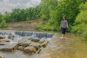 A woman in casual clothing walks along the edge of Pillsbury Crossing, a stone-lined creek with a small waterfall, surrounded by lush green trees and vegetation on a sunny KS day.
