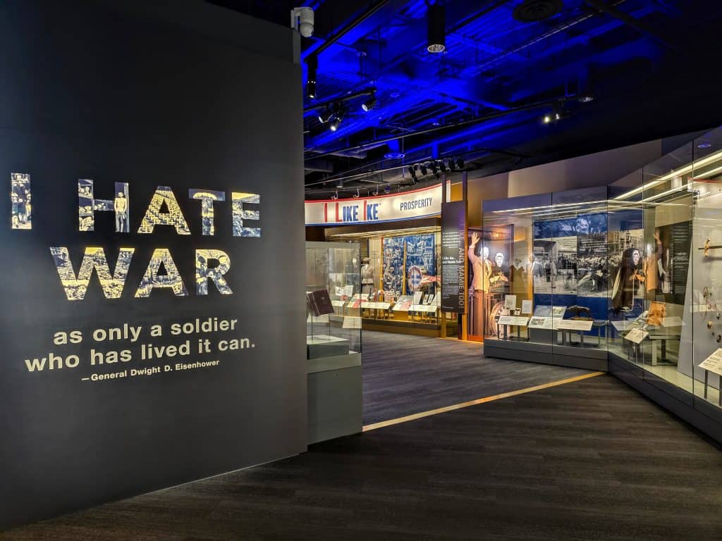 An exhibition hall in the Presidential Library of Dwight D. Eisenhower, located in Abilene, KS, features a quote by General Dwight D. Eisenhower: "I HATE WAR as only a soldier who has lived it can." The hall boasts displays on both sides and prominent blue lighting on the ceiling. A sign in the background reads "I LIKE IKE PRESIDENT.