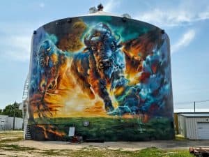 A large cylindrical structure in Clay Center, KS, is adorned with the vibrant Prairie Thunder Mural depicting two bison. The bison appear dynamic, one in the foreground and one in the background, surrounded by intense, colorful swirls of blue, green, and orange. A ladder is attached to the side.