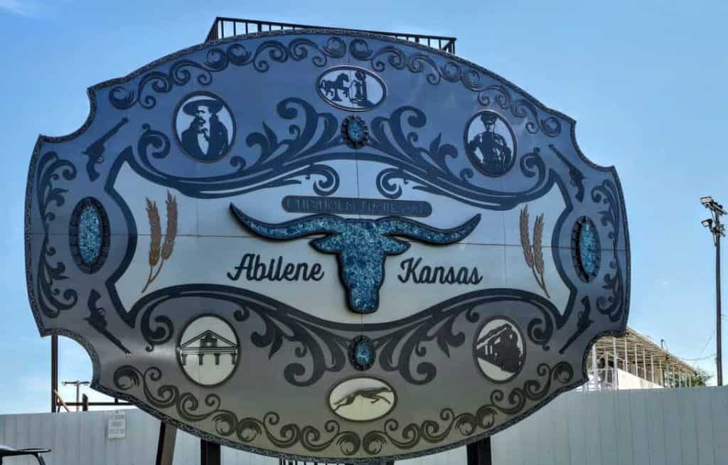 A large, ornate belt buckle replica stands on display in Abilene, Kansas. It features a blue longhorn in the center, surrounded by various detailed designs. Below the buckle, a sign reads "Discover the World’s Largest Belt Buckle." A utility vehicle is parked nearby.