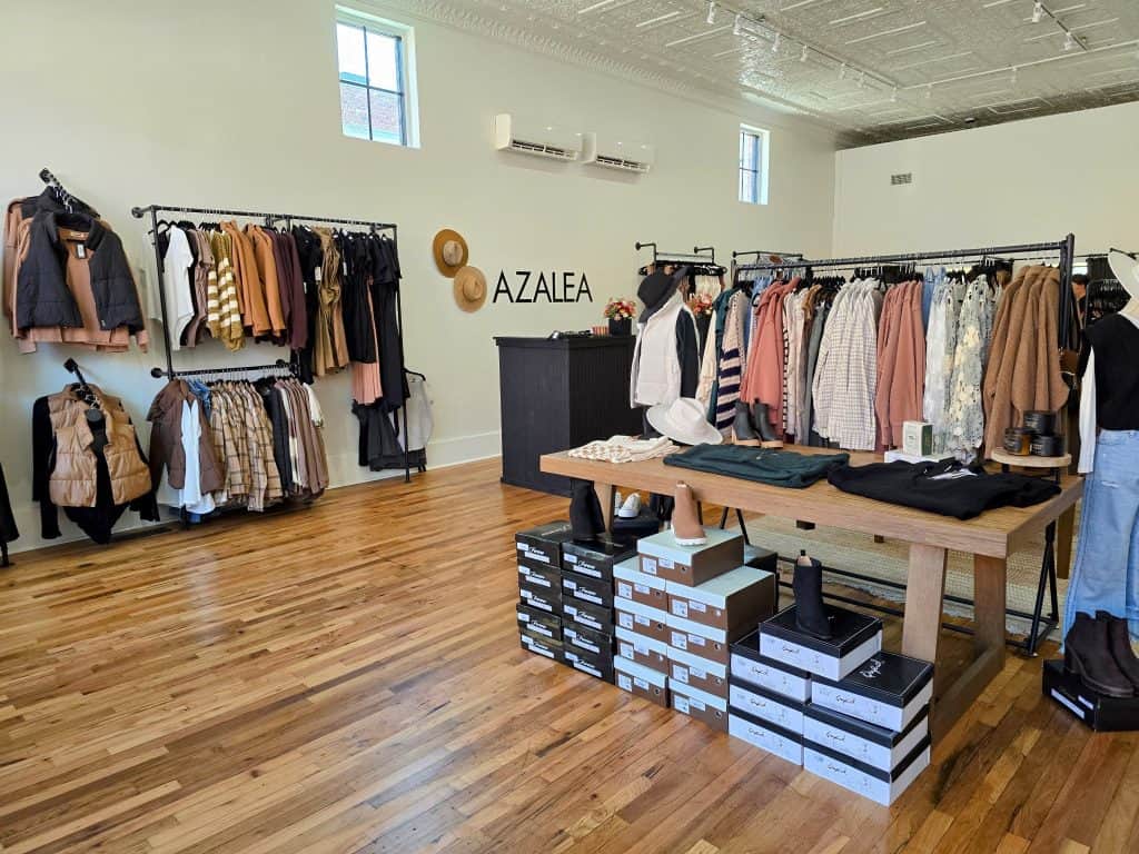A chic boutique with wooden floors displaying clothing on racks and shelves. The display at Azalea Avenue Boutique includes various shirts, jackets, and outerwear. Shoe boxes are stacked under a wooden table with accessories. The name "Azalea" is visible on the back wall, capturing the essence of KS style.