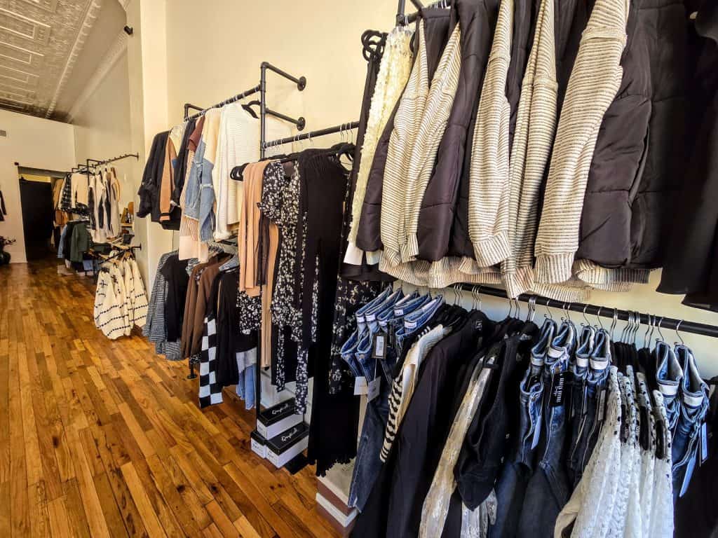A well-organized clothing store, Azalea Avenue Boutique, displays various garments on black metal racks against cream walls. The clothes include jackets, sweaters, shirts, and jeans. The wooden floor adds a warm touch to the neatly arranged store in KS.