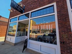 A brick building with large windows showcasing a clothing boutique named "Azalea Avenue Boutique" in Hays, KS. The storefront has white-framed windows, a black hanging sign above, and a sandwich board on the sidewalk. Clothing racks are visible through the window.