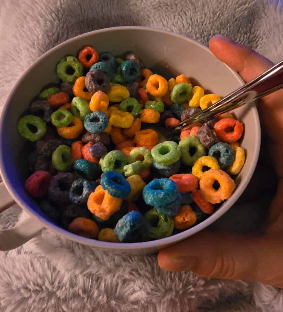 A hand holds a white bowl filled with colorful, loop-shaped cereal and milk, with a spoon resting inside. The person, likely tired from chemotherapy, is seated on or covered with a fluffy, gray blanket.