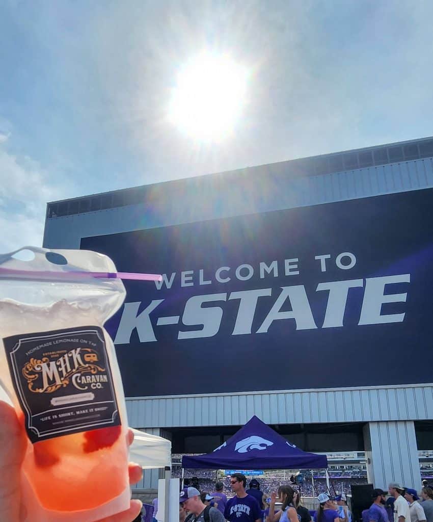 A person holds a drink in front of a large "Welcome to K-State" sign under the bright sun. The festive atmosphere of the Manhattan campus buzzes with people, tents, and the university's logo, reminiscent of an MHK Caravan celebration.