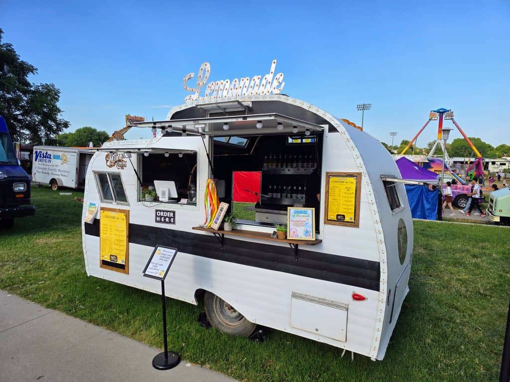 A vintage-style white and black lemonade trailer at the fair, marked as an MHK Caravan, with a bright "Lemonade" sign on top. The window is open, displaying a menu and drinks. Colorful tents and a Ferris wheel adorn the backdrop under Manhattan's clear blue KS sky.