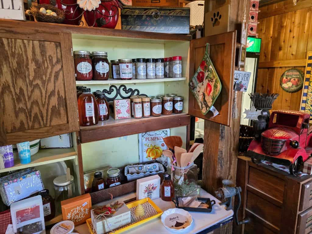 A cozy country store interior at Cider Hill features wooden shelves adorned with jars of homemade preserves, a vintage coffee grinder, and seasonal decorations. A cabinet door is open, revealing more jars and a decorative dish towel, evoking the charm of a family orchard near Kansas City.