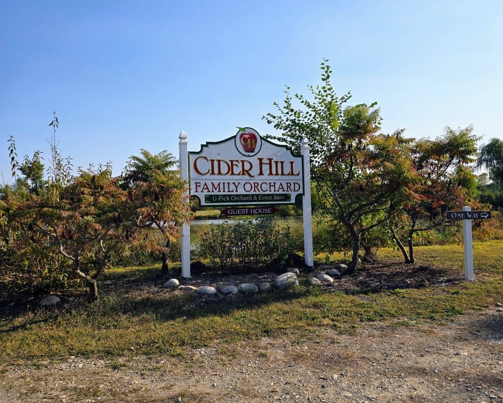 The sign for "Cider Hill Family Orchard" is nestled among trees and grass, hinting at its Kansas City charm. It mentions a guest house and highlights its U-Pick orchard along with an event barn. A smaller sign directs visitors to the right with "One Way.