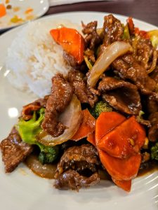 At Melanie's Kitchen in Hays, KS, enjoy a plate of stir-fried beef with onions, carrots, and broccoli served alongside a mound of white rice on a pristine white plate.