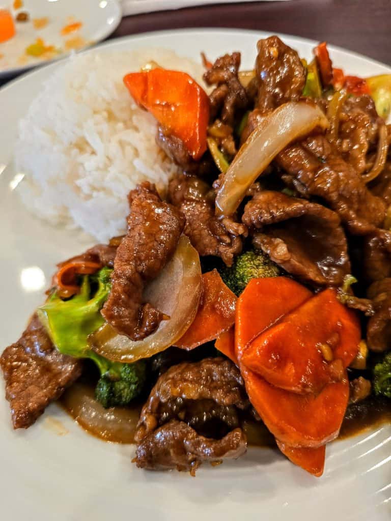 At Melanie's Kitchen in Hays, KS, enjoy a plate of stir-fried beef with onions, carrots, and broccoli served alongside a mound of white rice on a pristine white plate.