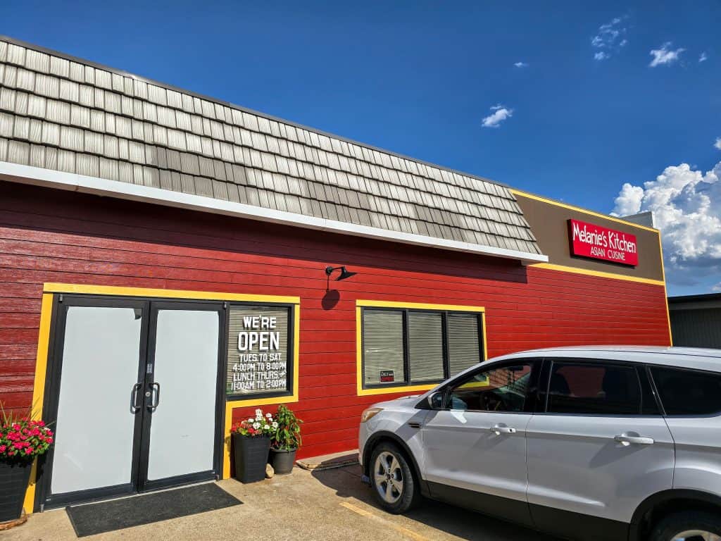 A red restaurant building with a sign reading "Melanie's Kitchen Asian Cuisine" stands proudly under the bright blue Hays sky. A white SUV is parked outside, and the entrance welcomes guests with a "We're Open" sign displaying business hours.