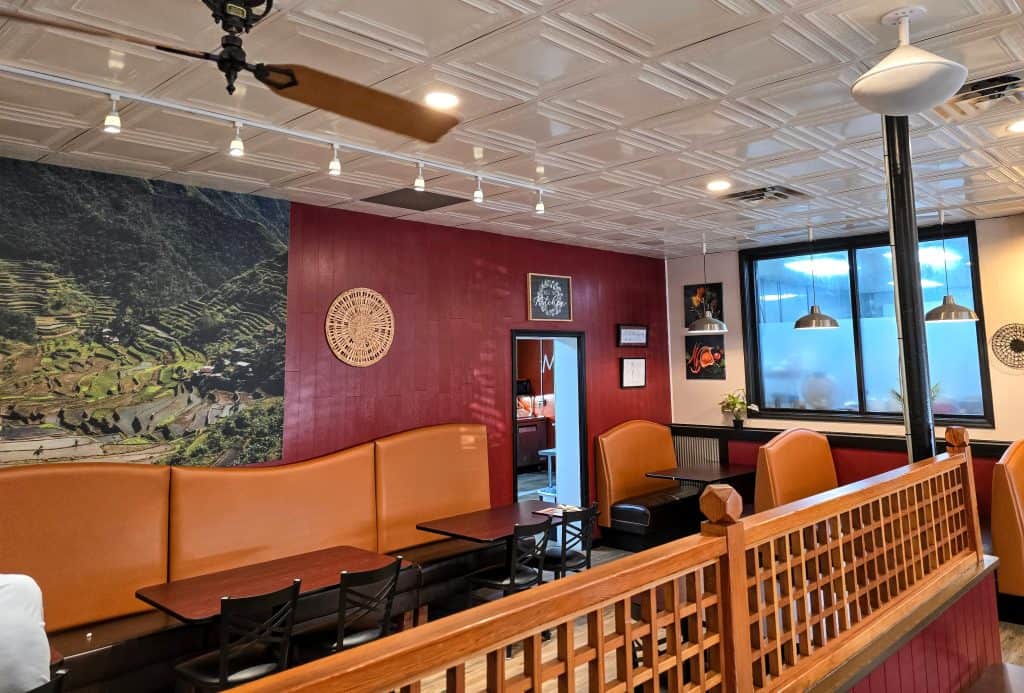 Melanie's Kitchen offers a cozy ambiance with red walls, white ceiling tiles, and wooden decor. Located in Hays, it features booth seating and tables lining the room. A mural of rice terraces and a decorative plate enhance the vibe, while ceiling fans and pendant lights provide lighting.