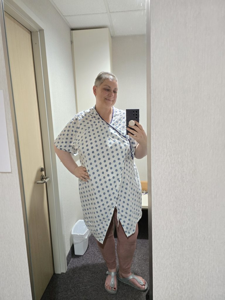 A person with short hair takes a selfie in a mirror, capturing their hospital gown with a polka dot pattern over pink pants and sandals. The setting, indicative of their chemo journey, includes a door and waste bin visible in the background.