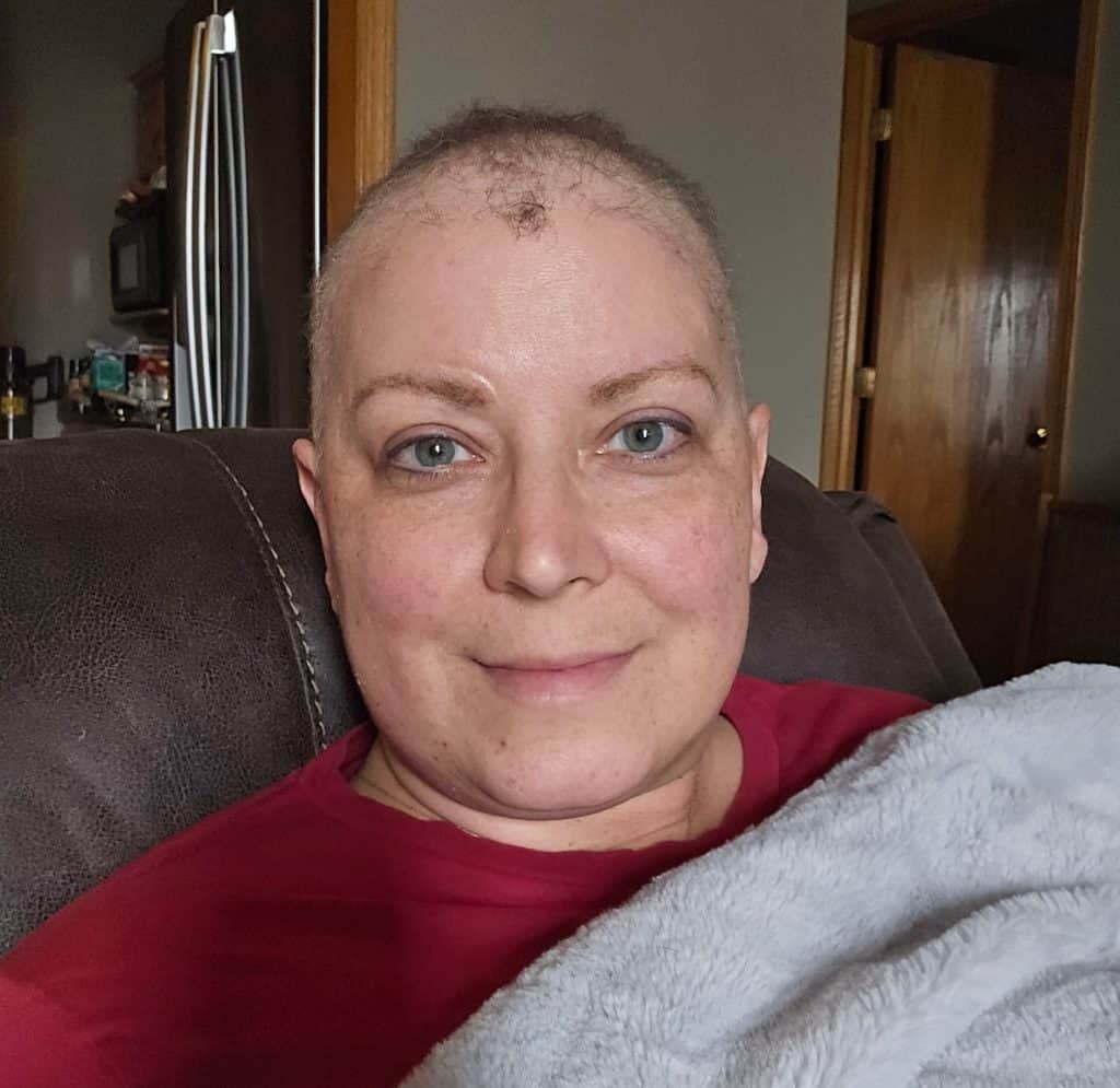 A person with short hair and blue eyes, dressed in a red top, sits on a dark couch partially covered by a gray blanket. They look directly at the camera with a neutral expression, perhaps contemplating their new chemo journey. The background showcases a wooden door and kitchen items.