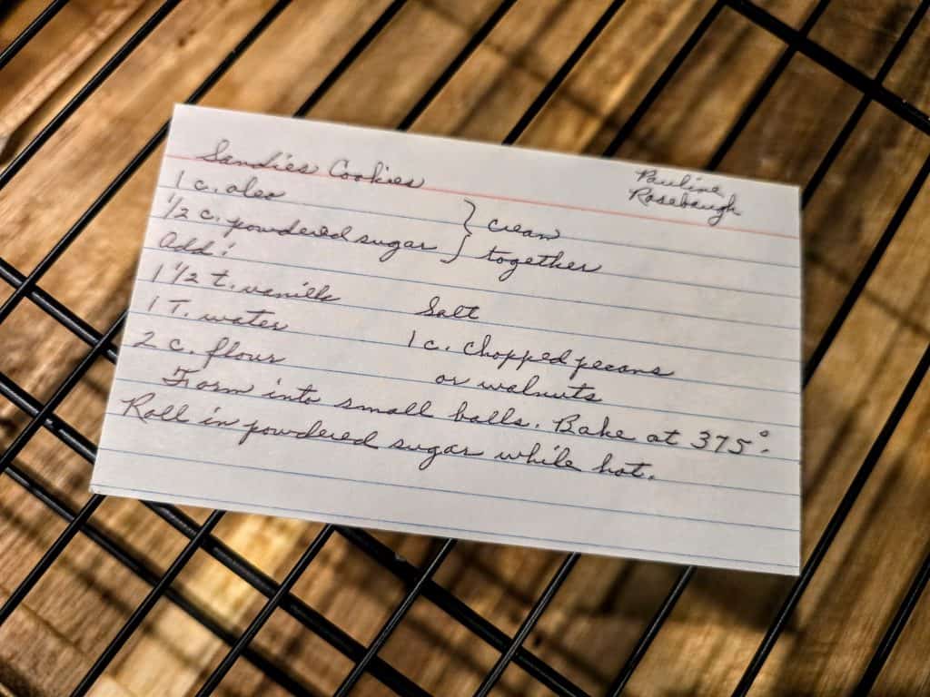 A handwritten recipe card on a wire rack, titled "Grandma Eva's Sandies Cookies," lists ingredients like powdered sugar, flour, vanilla, salt, pecans, and butter. It includes instructions for mixing and baking at 375°F.