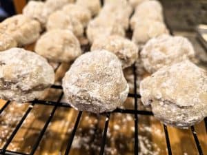Close-up of Grandma Eva's Sandies, powdered sugar-coated cookies cooling on a black wire rack, with a wooden surface underneath. The round, textured sandies showcase their delightful homemade appearance.