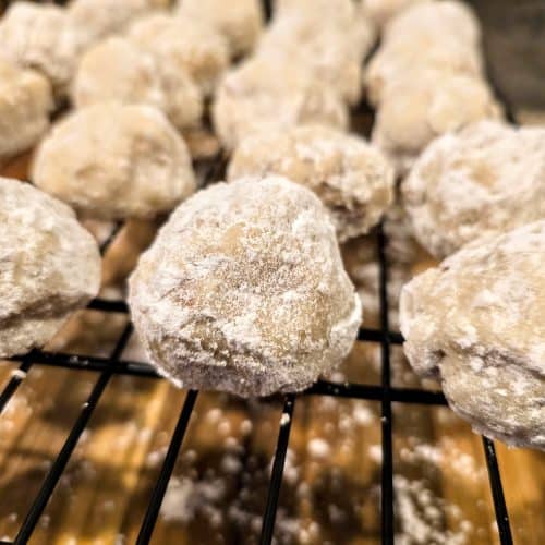 Close-up of Grandma Eva's Sandies, powdered sugar-coated cookies cooling on a black wire rack, with a wooden surface underneath. The round, textured sandies showcase their delightful homemade appearance.