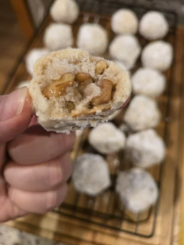 A close-up of a hand holding one of Grandma Eva's Sandies, a bite-sized cookie with a nut filling, showcasing its textured dough exterior and walnuts inside. More Sandies are cooling on a wire rack in the background.