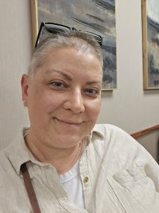 With short hair and glasses perched over their head, a person smiles warmly at the camera. They are wearing a light-colored shirt and sitting indoors, with abstract art radiating vibrant colors on the wall behind them.