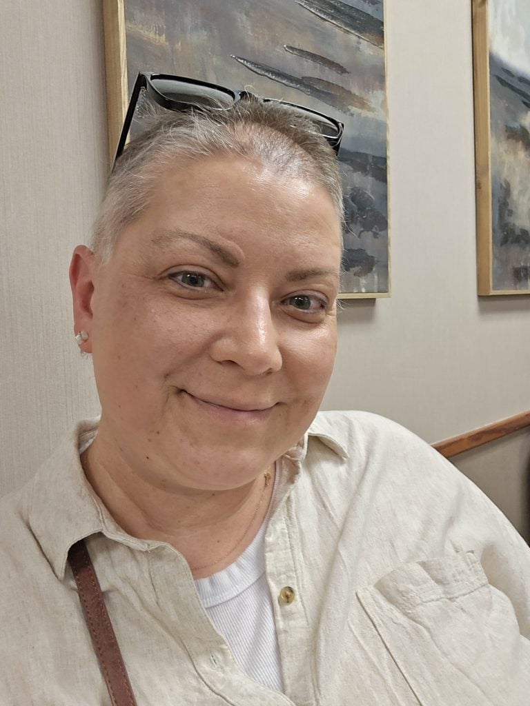 With short hair and glasses perched over their head, a person smiles warmly at the camera. They are wearing a light-colored shirt and sitting indoors, with abstract art radiating vibrant colors on the wall behind them.