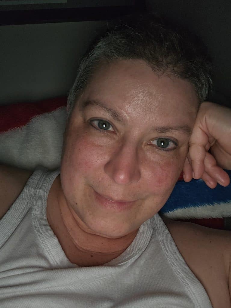 A person with short hair is lying down, resting their head on their hand. They're wearing a white tank top, looking directly at the camera with a slight smile. The background is dark with an overtone of mystery, and a colorful blanket is partially visible.