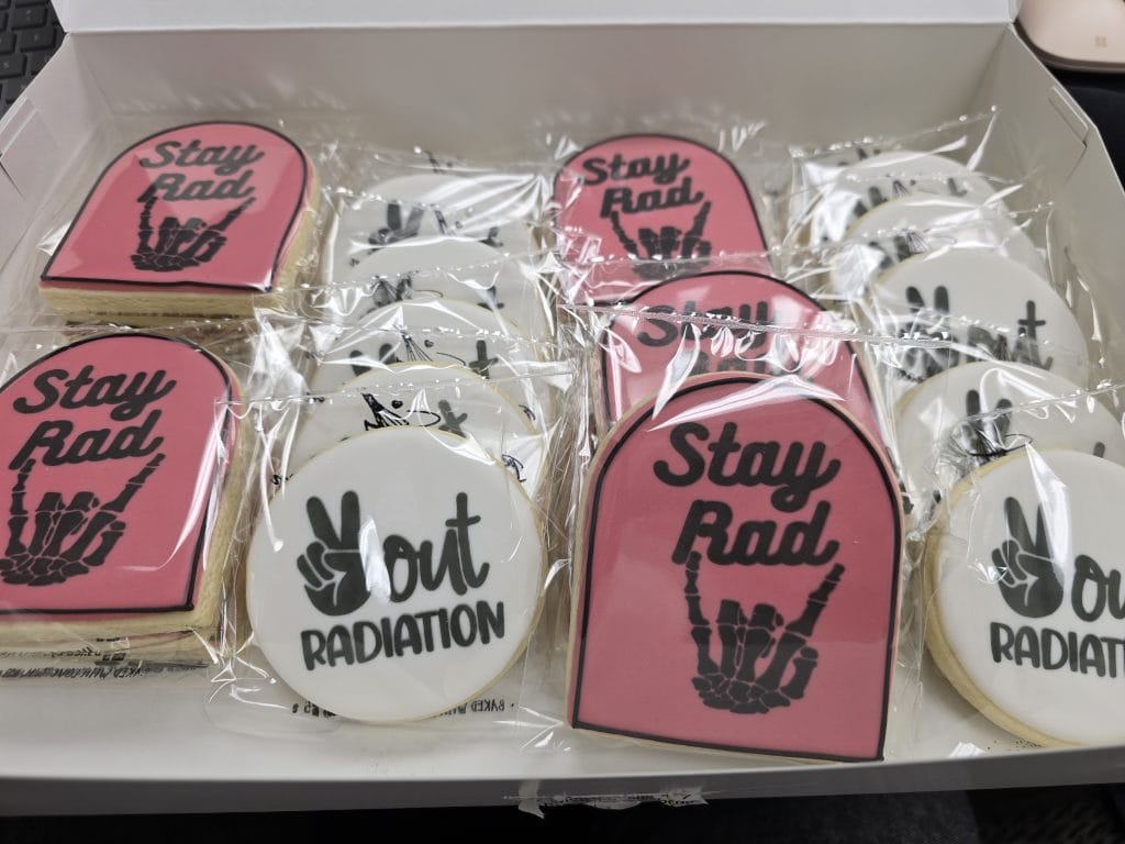 A box of cookies wrapped in plastic. Some cookies feature a pink background with "Stay Rad" and a skeleton hand symbol, subtly echoing breast cancer awareness. Others are white with "Yout Radiation" and a peace sign, adding a playful twist.