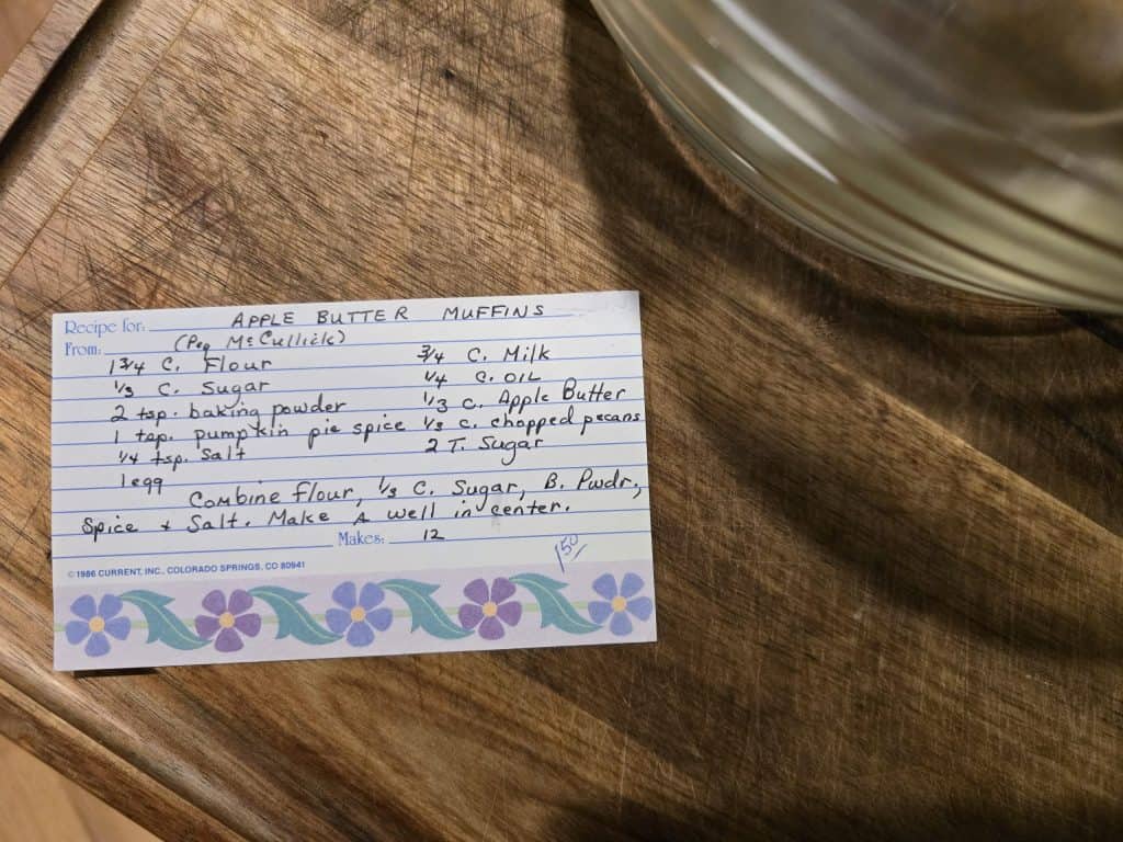 A handwritten recipe card titled "Grandma's Apple Butter Muffins" rests on a wooden surface. It showcases ingredients and instructions, framed by a decorative floral border on the left. A glimpse of a mixing bowl peeks out from the bottom right.