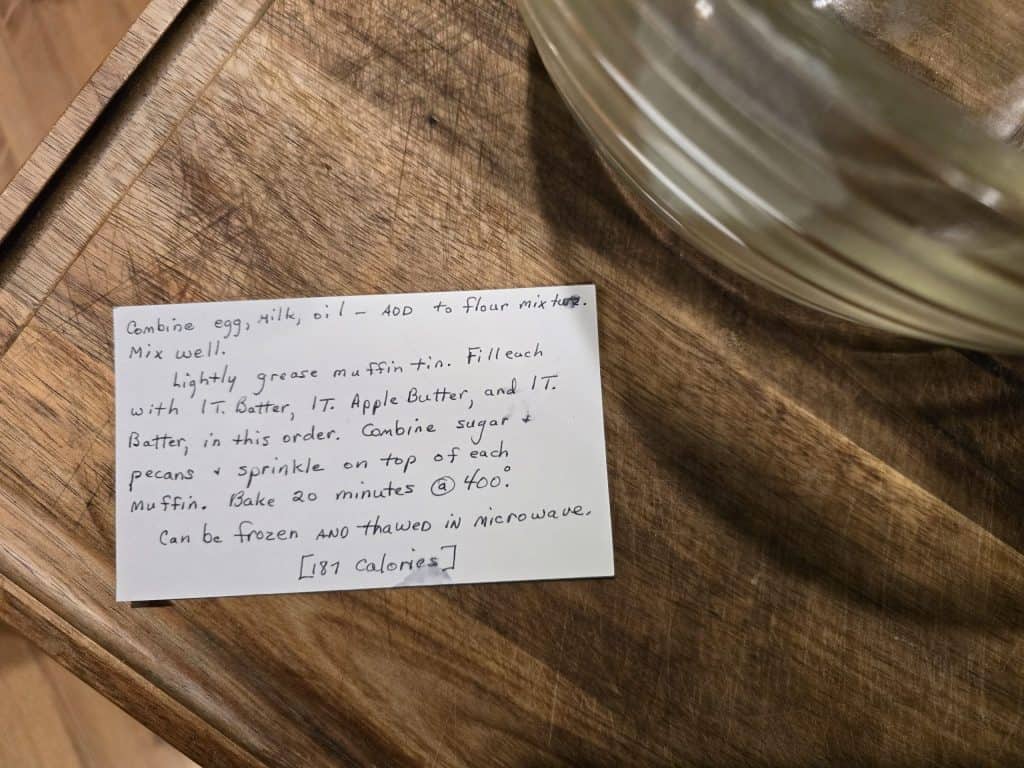 A handwritten recipe card on a wooden surface, titled "Oatmeal cups 101," features ingredients like mashed banana and oats. A note suggests adjustments for flavor and texture, reminiscent of Grandma's Muffins. A glass bowl is partially visible.