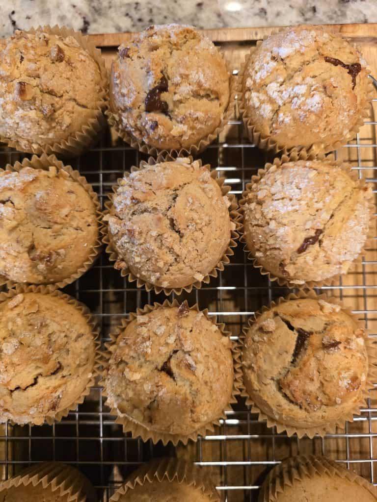 A close-up of twelve freshly baked muffins with a golden-brown crust, arranged in rows on a cooling rack. Some Grandma's muffins reveal chocolate chip pieces and a light dusting of sugar on top, with hints of apple butter wafting through the air. The background features a granite countertop.