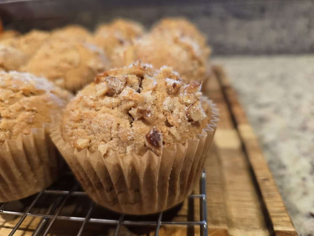 Close-up of freshly baked apple butter muffins with a crumbly, nutty topping, sitting on a cooling rack. The muffins are in brown paper liners, and the background shows a speckled countertop reminiscent of grandma's kitchen charm.