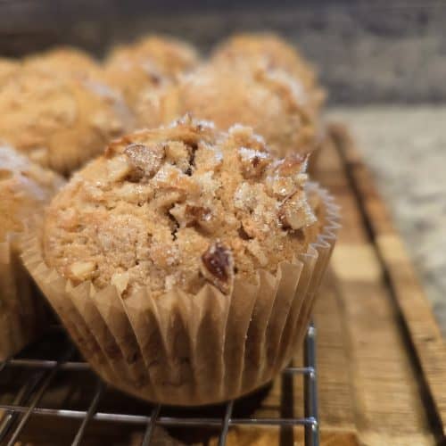 Close-up of freshly baked apple butter muffins with a crumbly, nutty topping, sitting on a cooling rack. The muffins are in brown paper liners, and the background shows a speckled countertop reminiscent of grandma's kitchen charm.