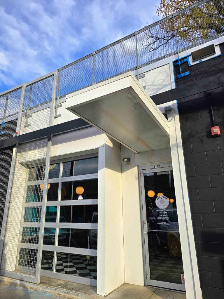 A modern building entrance in Manhattan boasts a glass door adorned with Full Circle white decals and a garage-style window. The door opens to the sidewalk, sheltered by an overhang, while a mesh fence lines the rooftop. Above, the KS sky is partly cloudy.