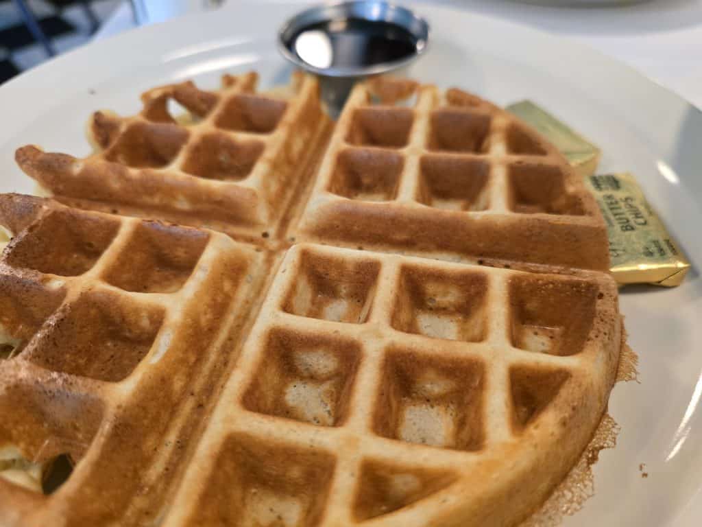 A close-up of a golden-brown waffle on a white plate, reminiscent of breakfast in Manhattan, KS. Accompanied by syrup and two packets of butter, the waffle is divided into quarters, highlighting its crisp texture—a warm start that brings things Full Circle.