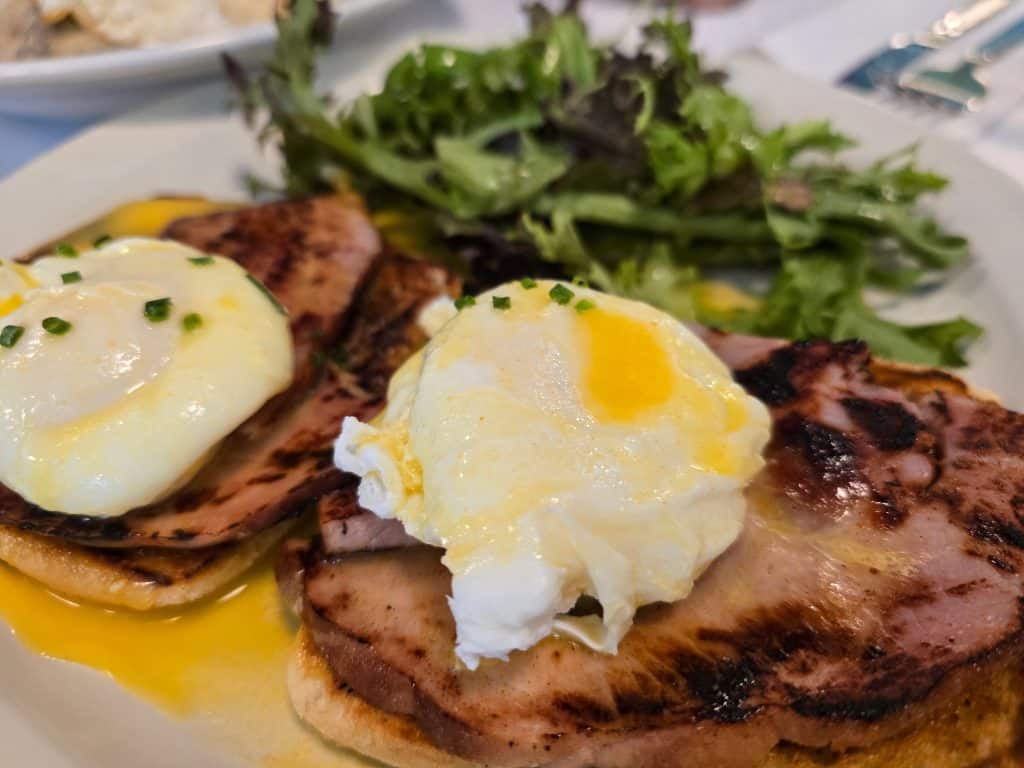 A plate of eggs Benedict with poached eggs and ham on an English muffin, topped with hollandaise sauce and sprinkled with chives. Served Full Circle style in Manhattan KS, this dish includes a side of mixed greens for a perfect finish.