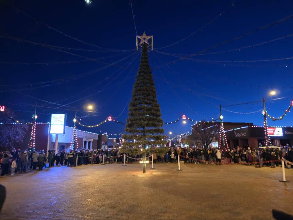 A tall, conical Christmas tree adorned with twinkling lights stands majestically in the heart of WaKeeney, known as the "Christmas City of the High Plains." Crowds gather around while vibrant string lights illuminate the evening sky.