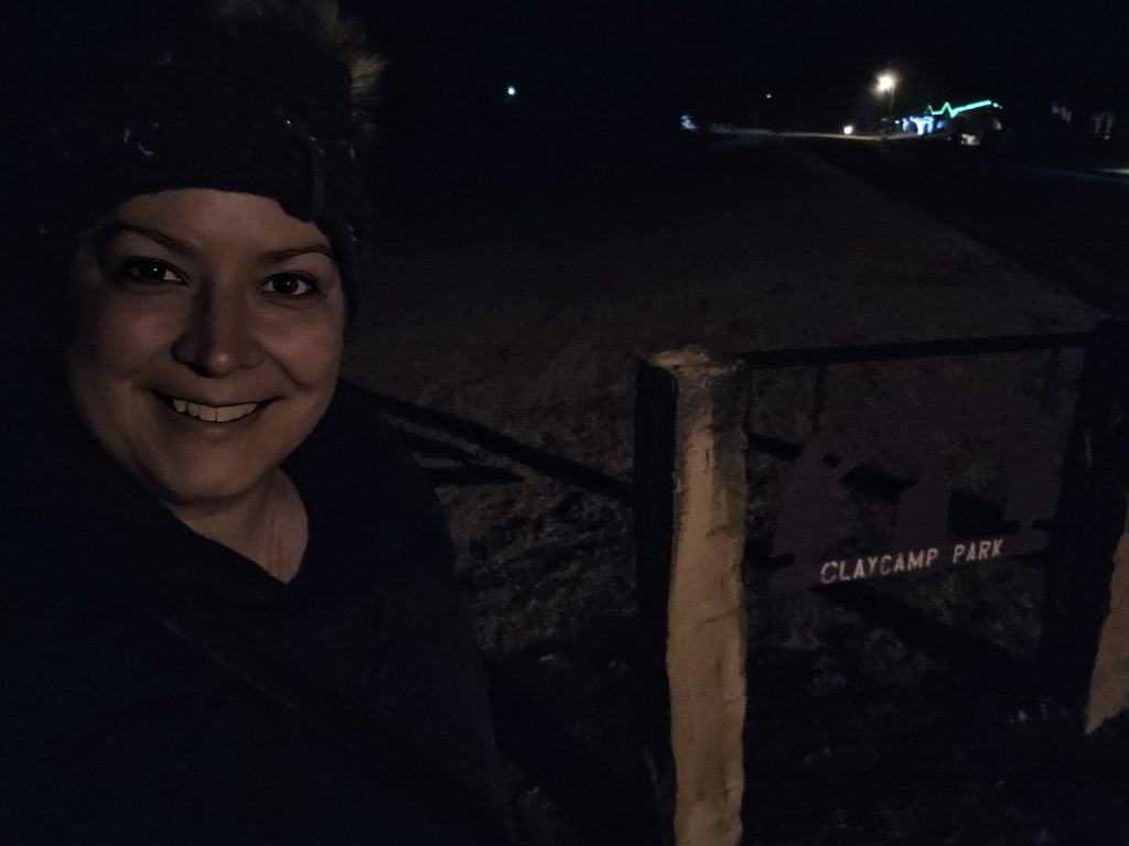 A person is smiling in front of a sign that reads "Claycamp Park" at night. The dimly lit area, located in the heart of WaKeeney, KS—often called the Christmas City—has faint lights twinkling in the distant High Plains background.