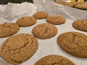 Freshly baked molasses cookies on a parchment-lined baking sheet. The cookies are golden brown, reminiscent of Grandma's Cookies, with a textured surface suggesting a soft and chewy interior.