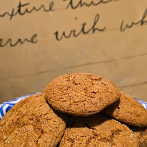 A plate of homemade molasses cookies stacked in a pile, with a handwritten recipe on brown paper in the background. The cookies are golden brown with a slightly cracked surface, reminiscent of Grandma’s classic treats.