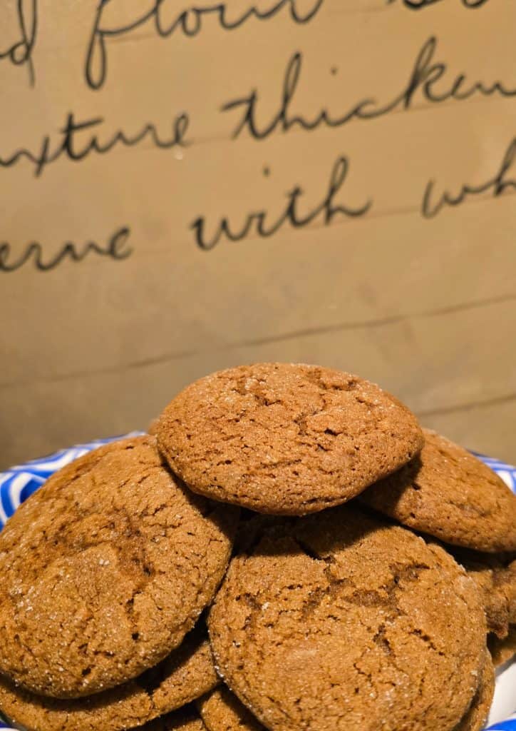 A plate of homemade molasses cookies stacked in a pile, with a handwritten recipe on brown paper in the background. The cookies are golden brown with a slightly cracked surface, reminiscent of Grandma’s classic treats.