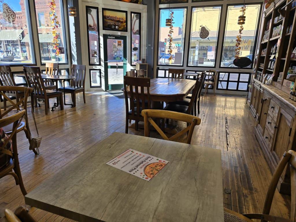 A cozy café in Hays, KS, with wooden floors, tables, and chairs. Sunlight streams through large windows adorned with hanging decorations. A menu at Paisley Pear lies on a table in the foreground, and various items are attractively displayed on the shelves.