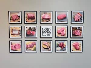 A collage of framed images showcases various cuts of beef, including steaks and ground beef, elegantly arranged around a central "1880 Beef" sign. Each cut is beautifully displayed on wooden boards, garnished with herbs and spices&mdash;a true homage to Manhattan KS's rich beef tradition.