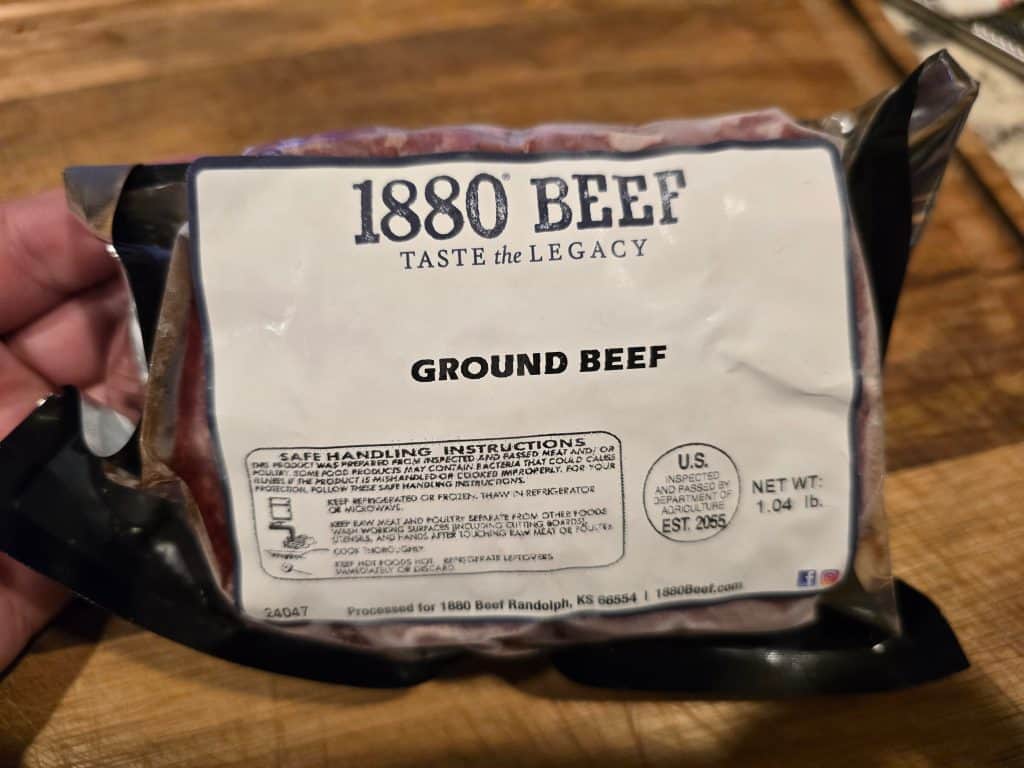 A hand holds a package of 1880 Beef, sourced from Manhattan, KS, labeled "GROUND BEEF," weighing 1.04 pounds. The label features safe handling instructions and a brand description. All of this is set against a rustic wooden backdrop.