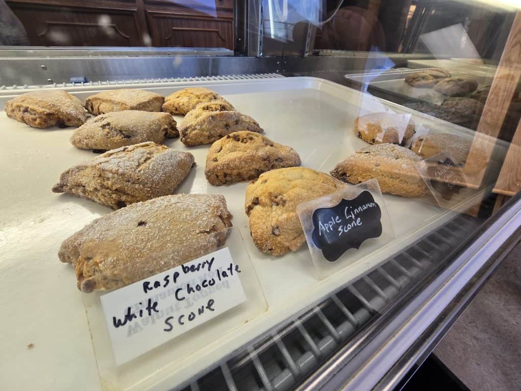 A display case at Augustine's Bakery showcases raspberry white chocolate scones on the left and apple cinnamon scones on the right. A small chalkboard sign identifies the latter. Both varieties sit invitingly on a pristine white tray, tempting everyone in Hays to indulge.