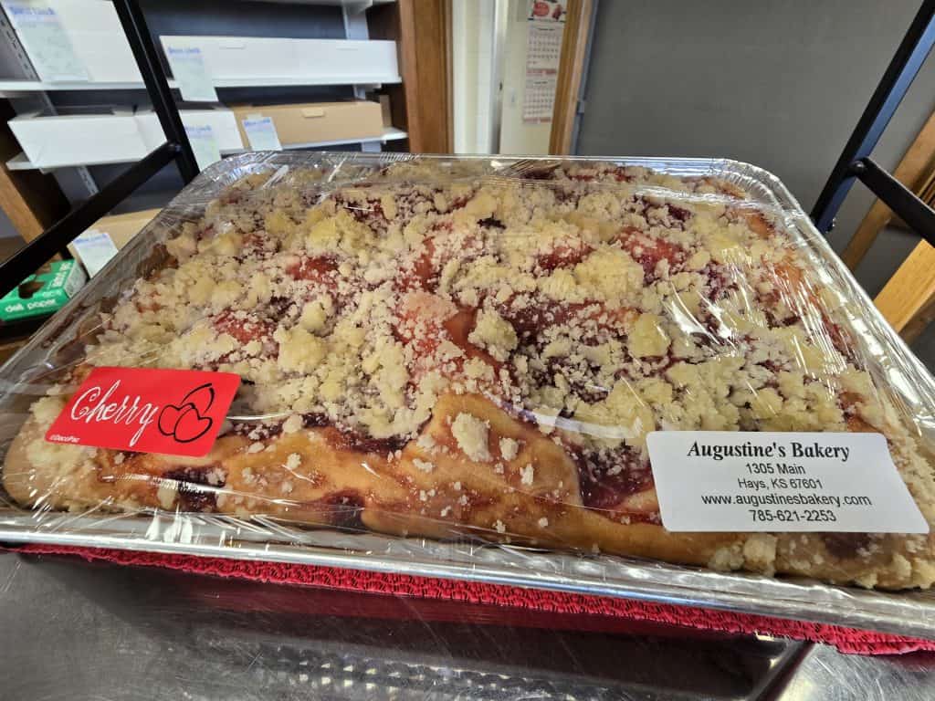 A large tray of cherry pastry from Augustine's Bakery in Hays, KS, is covered in clear wrap. The crumbly topping tempts the taste buds. A red label with a cherry graphic reads "Cherry." The bakery's contact information is on a sticker in the corner.