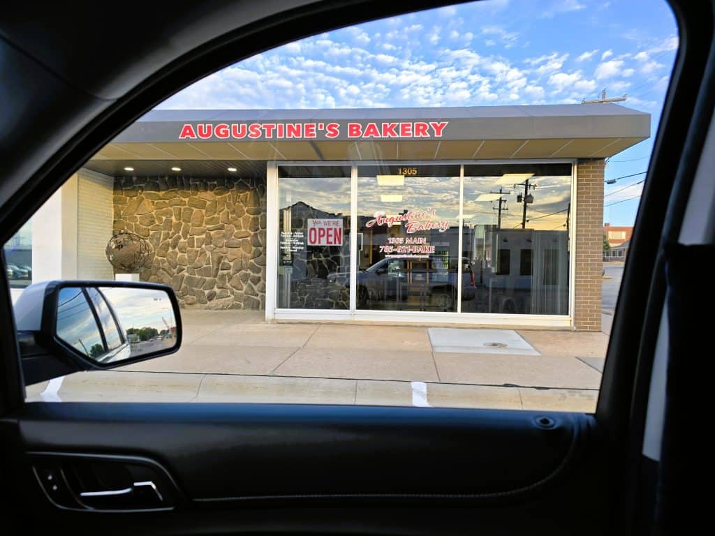 Through the car window, you see Augustine's Bakery in Hays, KS, with its large windows and stone wall section. A glowing red "OPEN" sign beckons beneath the bold red letters of its name. Reflections dance in the glass, inviting you to explore inside.