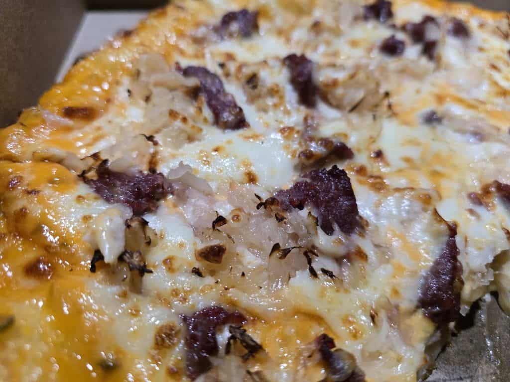 This close-up of Lomato's Pizza showcases a golden-brown crust topped with melted cheese, caramelized onions, and savory slices of beef or another type of meat, all served in a classic cardboard box.