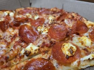 A close-up of a freshly baked Lomato's pepperoni pizza in a cardboard box. The pizza features browned, crispy pepperoni slices and melted cheese on a golden crust, representing the delicious quality Lomato's is known for.