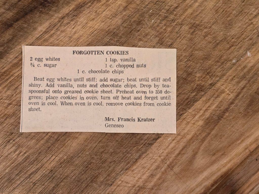 A recipe card titled "Grandma's Forgotten Cookies" on a wooden surface. Ingredients include egg whites, sugar, vanilla, nuts, and chocolate chips. Instructions detail mixing, dropping spoonfuls on a sheet, baking at 350°F, then cooling in an off oven.