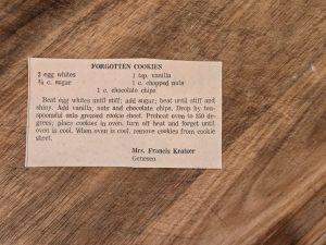 A recipe card titled "Grandma's Forgotten Cookies" on a wooden surface. Ingredients include egg whites, sugar, vanilla, nuts, and chocolate chips. Instructions detail mixing, dropping spoonfuls on a sheet, baking at 350&deg;F, then cooling in an off oven.